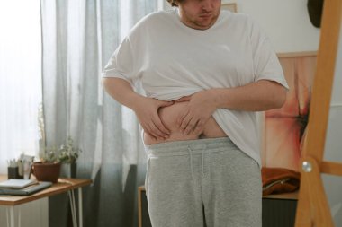 Perdelerden süzülen doğal ışıkla normal bir evde karnını muayene eden bir adam. Odada küçük bir masa ve iç mekan bitkileriyle rahat bir dekor var.