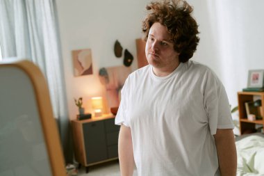 Rahat yatak odasında duran kıvırcık saçlı bir adamın portresi, rahat bir ifadeyle kameraya bakıyor. Beyaz gömlekli, aydınlık, basit dekorlu bir odada.