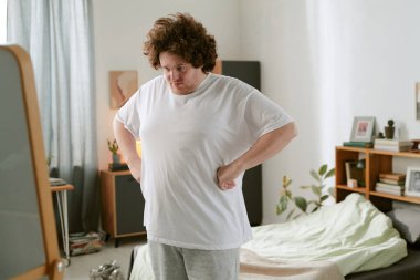 Kıvırcık saçlı bir adam, parlak bir odada, dağınık yatağı ve sabah rutinini gösteren çeşitli kişisel eşyalarıyla kendini yansıtıyor.