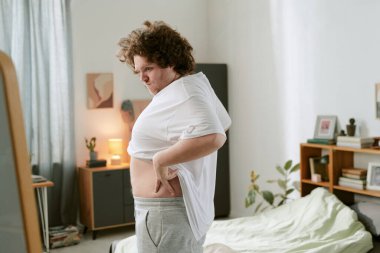 Sıcak bir yatak odasında beyaz bir tişört ayarlayan bir birey. Odada bir yatak, kitaplık ve sıcak ve davetkar bir atmosfer yaratan kapalı bitkiler var.