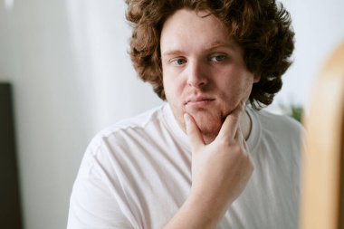 Saçları kıvırcık, çenesine dokunan, derin düşüncelere dalmış genç beyaz adamın portresi. Beyaz gömlek, iç mekan ayarı, tarafsız arka plan, hafif ışıklandırma.