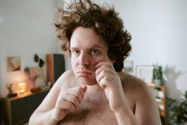 Kıvırcık saçlı ve ciddi ifadeli bir adamın portresi, rahat bir oturma odasında, arka planda raflar ve bitkilerle boks yapmaya hazırlanıyor.