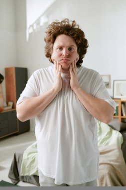 Gündelik giysiler giyen, modern bir dairede duran, yüzünü tutan ve dalgın görünen bir adamın portresi. Mobilya ve ev dekorasyonu arka planda görünür