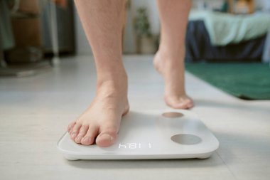 Dijital ölçekte ayakta duran ve ev ortamında görünen ağırlık ölçümleri yapan kişi. Arka planda ev eşyaları ve mobilyalar yer alıyor, sıcak atmosfer vurgulanıyor