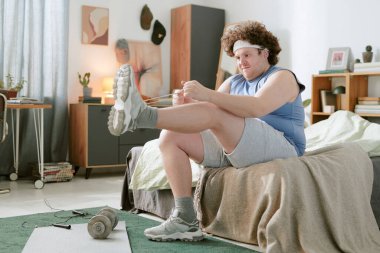 Genç, kıvırcık saçlı, yatakta oturan, bacaklarını esneten ve azimle egzersiz yapan beyaz bir adam. Ahşap mobilyalar ve ev dekorasyonu aksanıyla döşenmiş bir oda.