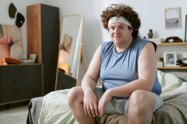 Saç bandı olan kıvırcık saçlı bir adamın portresi rahat bir pozla yatağın kenarına oturmuş, modern yatak odası dekoruyla çevrili, şifonyer ve raflar da dahil.