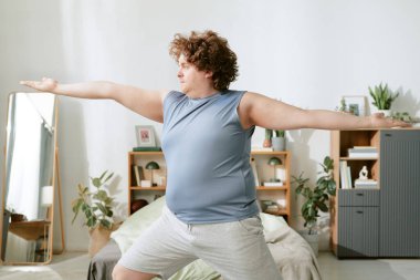 Modern oturma odasında yoga yapan bir adam raflar, bitkiler ve dekorla. Rahat bir kıyafet giymek ve açık bir alanda esneme egzersizi yapmak.