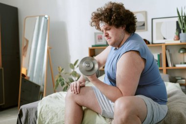 Adam oturma odasında otururken dambıl kullanarak ev egzersizi yapıyor. Rahat ev ortamında fiziksel uygunluğun korunmasına odaklanmış