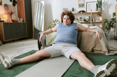 Modern yatak odasında bağdaş kurmuş, minimalist bir dekorla, mobilya ve bitkilerle çevrili kıvırcık saçlı, günlük kıyafetler ve spor ayakkabılar giyen bir adam.