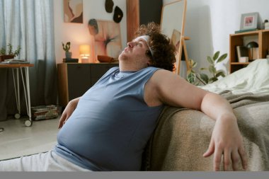 Sıcak, iyi dekore edilmiş spor salonunda yoğun egzersizden sonra yatakta dinlenen adam terli ve kolsuz tişört giyen yorgun bir ifadeyle yukarı bakıyor.