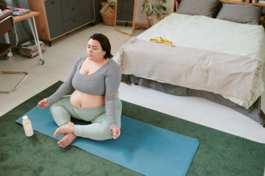 Mavi paspas üzerinde yoga ve meditasyon yapan bir kadın rahat bir odada arka planda kitaplık ve yakınlarda bir şişe su olan bir odada.