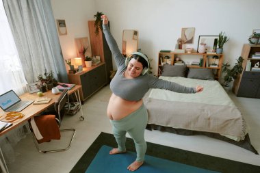 Yatak odasında kulaklıkla yoga egzersizi yapan bir kadın. Laptop, dokümanlar ve bitkilerle görülebilir bir rahatlama ortamı yaratıyor.