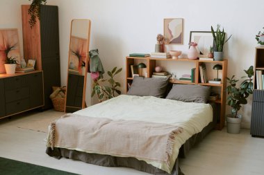 Ahşap mobilyalar, yeşil bitkiler ve rahat yatak örtüleriyle süslü şık bir yatak odası. Kitaplar ve sanat eserleriyle dolu bir oda, sıcak ve davetkar bir ortam sağlıyor.