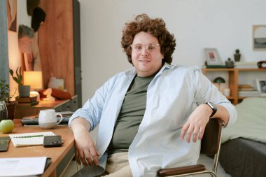 Gözlüklü bir adamın portresi rahat bir ofiste düzenli bir masada oturuyor. Bitkiler, kitaplar ve kişisel eşyalarla dekore edilmiş bir oda sıcak bir ortam yaratıyor.