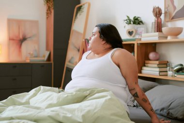 Yatakta sakin bir sabahtan zevk alan bir kadın rahat, aydınlık bir yatak odasında beyaz bir kolsuz bluz giyiyor. Arka plandaki ayna yatak odasının içini yansıtır