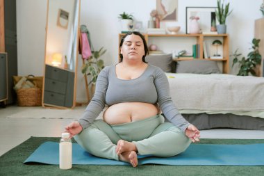 Hamile kadın yatak odasında meditasyon yapıyor. Etrafı sakinleştirici dekor ve hafif ışıklandırma ile çevrili, gözleri kapalı, yoga minderinde oturmuş su şişesi tutuyor.