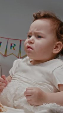 Kirli elleri olan küçük bir kızın dikey görüntüsü. Şık elbiseler giyiyor. Kremayla kaplanmış kaşıkları yalıyor ve doğum günü kutlamalarında balonlarla oynarken gülümsüyor.