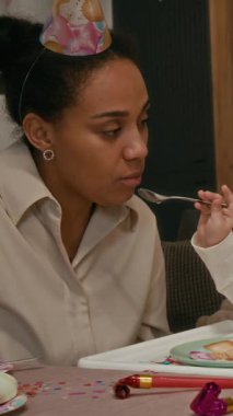 Doğum günü partisinde anne karnını pasta ile doyuran küçük kızın dikey görüntüsü, kızın yanında oturan baba.