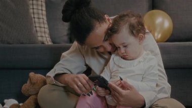 Oturma odasında yerde otururken akıllı telefonuyla oynayan küçük kızla konuşan genç bir kadın.
