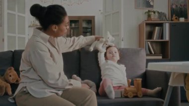 Genç anne, oturma odasında kanepede yan yana otururken küçük kızların kafasını yumuşak oyuncaklarla okşuyor.