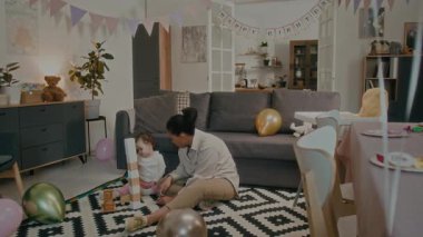 Küçük anne ve kızı tahta oyuncaklarla oynuyorlar. Kız, halı üzerinde oturmuş balonlarla süslenmiş akıllı telefon kullanan bir kızın fotoğrafını çekiyor.