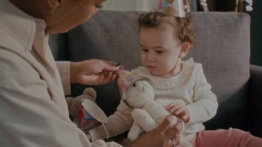 Küçük anne ve kız, oturma odasındaki kanepede birlikte oynarken lüks oyuncaklara parti şapkaları takıyorlar.