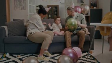 Neşeli çoklu etnik aileler kendilerine ve küçük kızlarına parti şapkaları takıp oturma odasındaki kanepede otururken renkli balonlarla oynuyorlar.