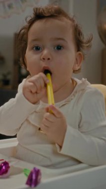 Kameraya poz veren ve evde beslenme koltuğunda otururken parti düdükleriyle oynayan iki yaşındaki pozitif kızın dikey portresi.