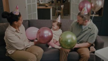 Doğum günü partisinde birlikte kanepede otururken parti şapkası takıp balonlarla oynayan pozitif ebeveynlerin ve bebek kızlarının yüksek açılı görüntüsü.