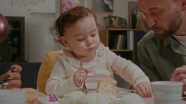 Genç ebeveynler, oturma odasındaki masada bayram yemeği yerken parti şapkası takan doğum günü kızına kaşıkla pasta yediriyorlar.