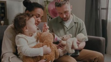 Bebek kız annesinin kucağında otururken oyuncak ayıyı tutuyor, baba doğum günü partisinde koltukta dinlenirken pelüş oyuncaklarla oynuyor.