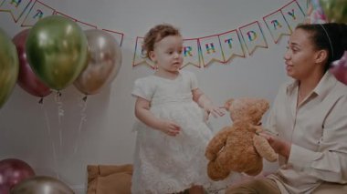 Doğum günü partisinde bayraklar ve balonlarla süslenmiş beyaz elbiseli bebeğe oyuncak ayıyı gösteren pozitif bir anne.