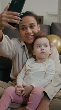 Neşeli annenin küçük kızıyla fotoğraf çektiği, evde dinlenirken altın balon çektiği dikey manzara.