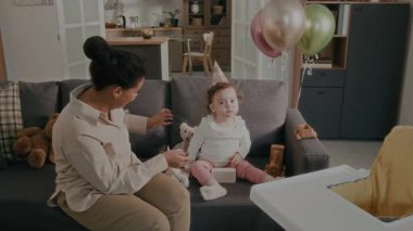 Genç anne, bebek kıza ve kendisine parti şapkası takıyor, sonra da koltukta akıllı telefon kullanarak onların selfie fotoğraflarını çekiyor.