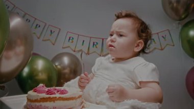 Beyaz elbiseli bebek, doğum günü partisinde kremalı pastayla beslenme koltuğunda otururken kaşık yalıyor ve renkli balonlar itiyor.