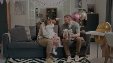Pozitif baba iki yumuşak oyuncakla oynuyor, küçük kız annesinin kucağında otururken onu izliyor oturma odasında doğum günü partisinde.
