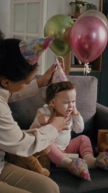 Annenin yaramaz bebek kızına parti şapkası takmaya çalışırken evde doğum günü kutlaması sırasında oyuncaklarla kanepeye oturmasının dikey görüntüsü.