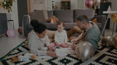 Çok ırklı aileler, doğum günü partisinde oturma odasındaki halının üzerinde işaretler kullanarak kelebek ve küçük kızın resimlerini boyuyor.