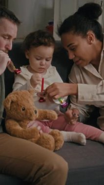 Evde kanepede otururken parti düdüğü çalan iki yaşındaki kız ve genç eşlerin mutlu bir aile fotoğrafı.