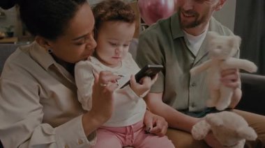Baba, anne dizlerinin üzerinde oturan ve aile fotoğrafı çekilirken ekrana dokunan küçük kız için akıllı telefonu açıyor.