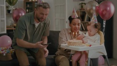 Yemek sandalyesinde oturan karısı ve kızıyla selfie çeken bir adam, doğum günü pastasında mumları söndüren ve el çırpan bir anne-baba.