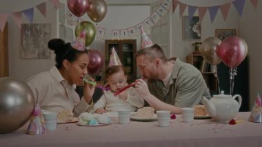 Mutlu ebeveynler parti düdüğü çalıyor, baba bebek kızlarının doğum gününü kutluyor oturma odasında balonlarla ve bayrak çizgileriyle süslenmiş bir tabak pastayla.