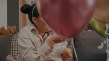 Neşeli anne, oturma odasında kanepede elma yiyen bebek kızıyla renkli balonlar itiyor.