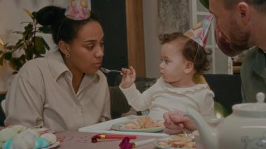 Küçük kız, doğum günü kutlaması sırasında babasının yanında otururken annesine pasta yediriyor.