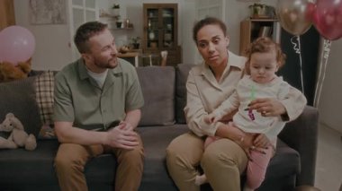 Doğum günü kutlaması sırasında annesinin kucağında oturan ve ağzında parti düdüğü olan bebeğe hediye paketi veren pozitif bir baba.