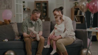 Babası oyuncak hayvanı, annesinin dizlerinin üstüne çöküp doğum günü kutlaması sırasında oyuncak koparan küçük kızına hediye kutusunu gösteriyordu.