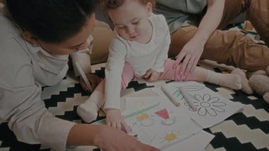 Anne, resimde kimin betimlediğini soran bebeği işaret ediyor. Sonra da oturma odasındaki halının üzerinde aile aktivitesi sırasında kızın babasından işaret alıyor.