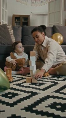 Tahta blokları olan mutlu anne kulesinin dikey görüntüsü, bebek iki yumuşak oyuncağı tutarken onu kırıyor.