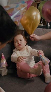 Farklı ailelerin renkli balonlarla oynamaları ve doğum günü partisinde koltukta oturan küçük kızla etkileşime girmeleri.