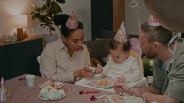 İki yaşındaki bir kız, doğum gününü şenlik masasında genç ebeveynleriyle kutluyor, evde pasta yiyor ve parti şapkalarında çay içiyor.